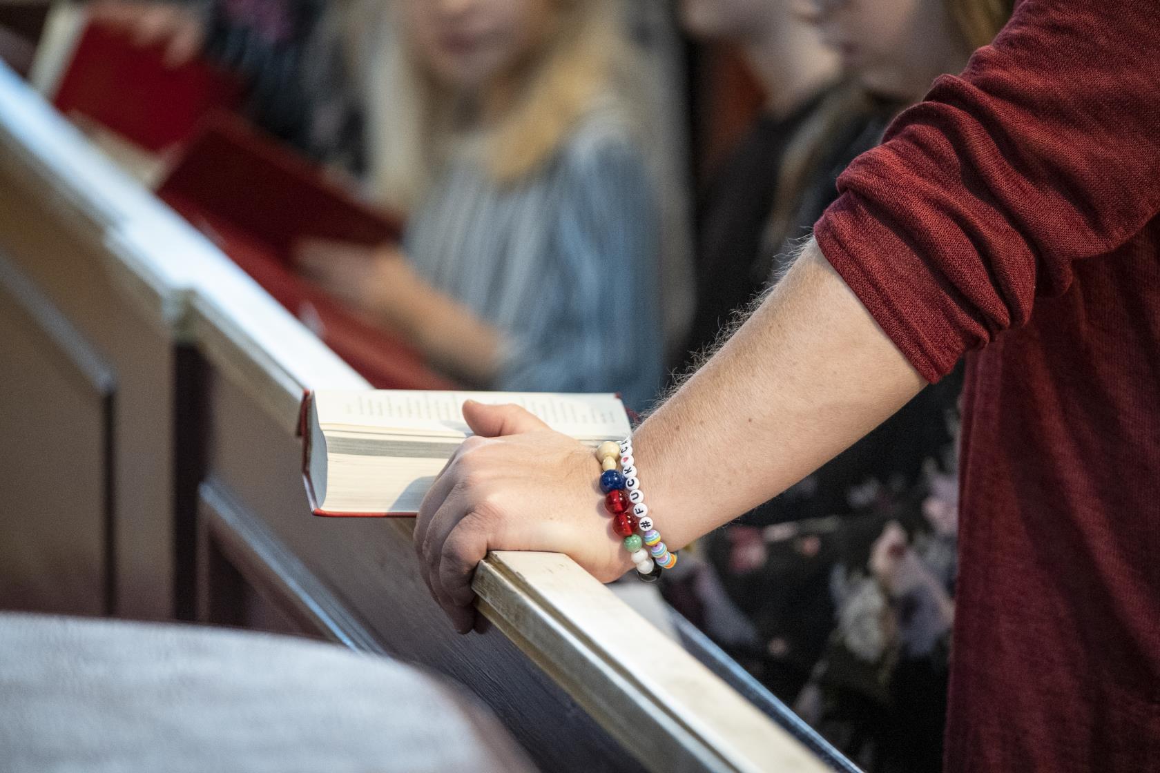 En person med ungdomliga armband stöder armen mot kyrkbänken, där en psalmbok ligger uppslagen.