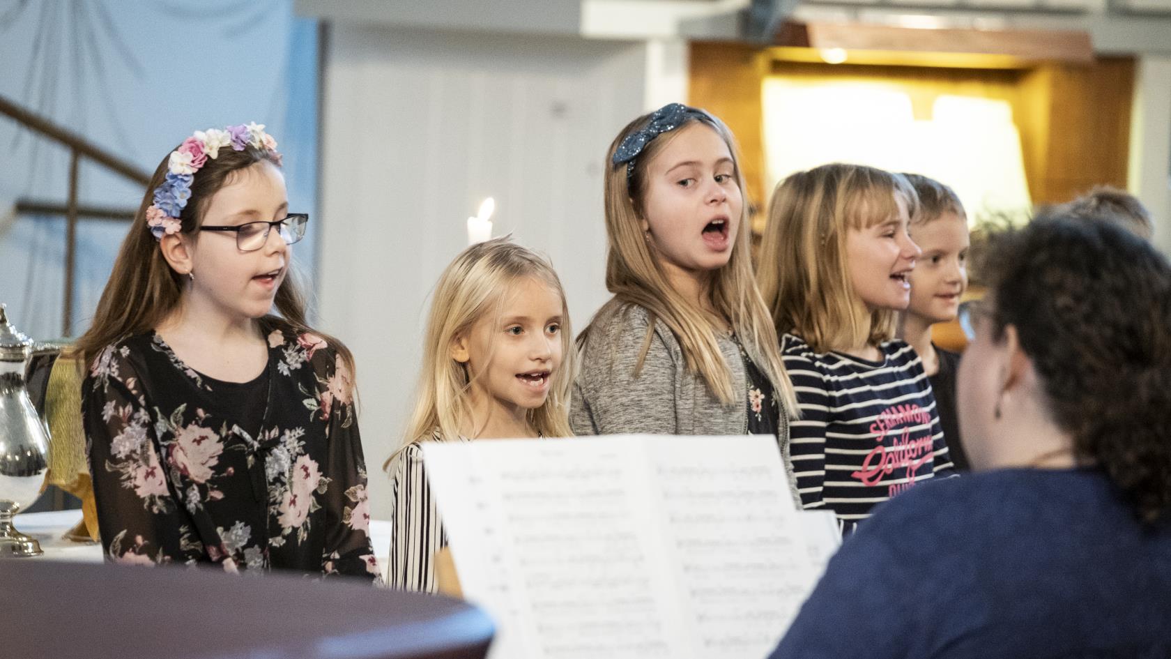 Några barn står bredvid varandra och sjunger framme i kyrkan. En kvinna kompar på piano.