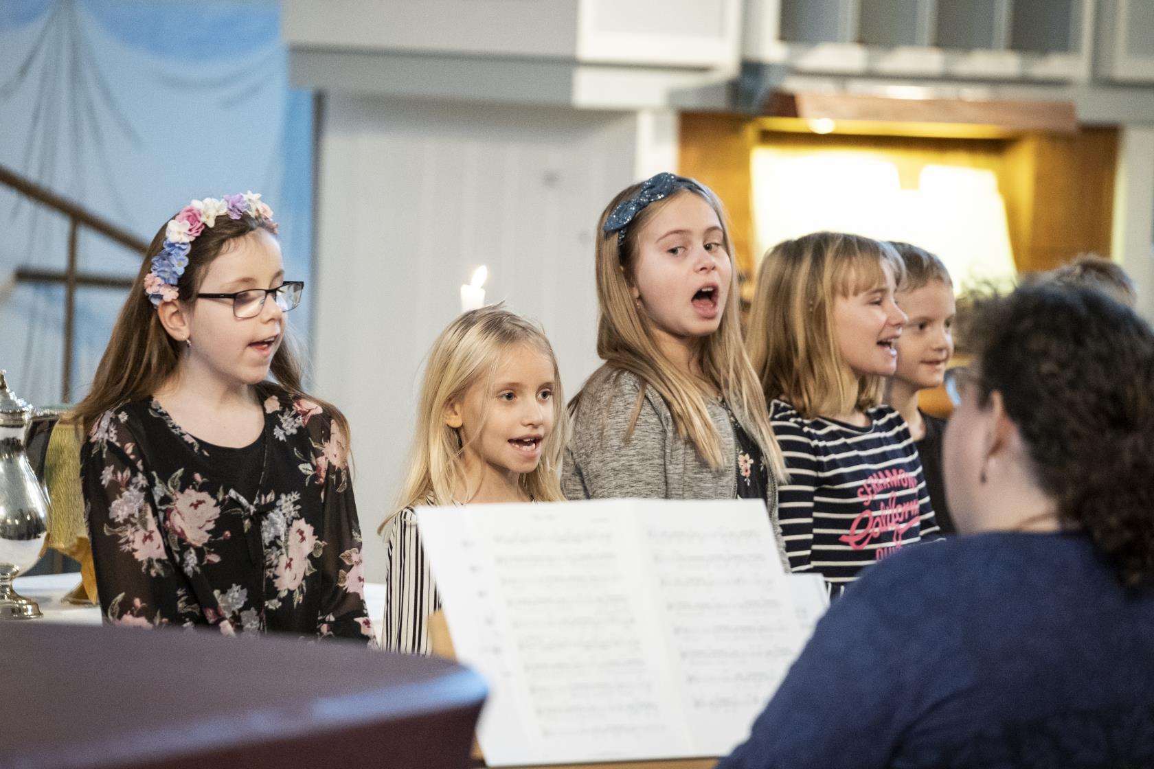 Några barn står bredvid varandra och sjunger framme i kyrkan. En kvinna kompar på piano.