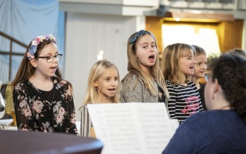 Några barn står bredvid varandra och sjunger framme i kyrkan. En kvinna kompar på piano.