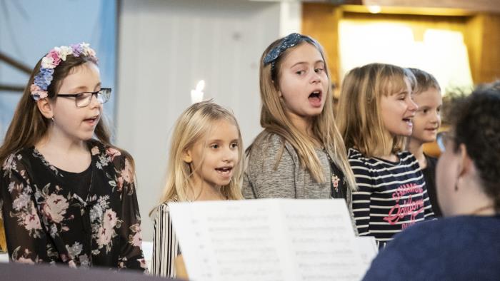 Några barn står bredvid varandra och sjunger framme i kyrkan. En kvinna kompar på piano.