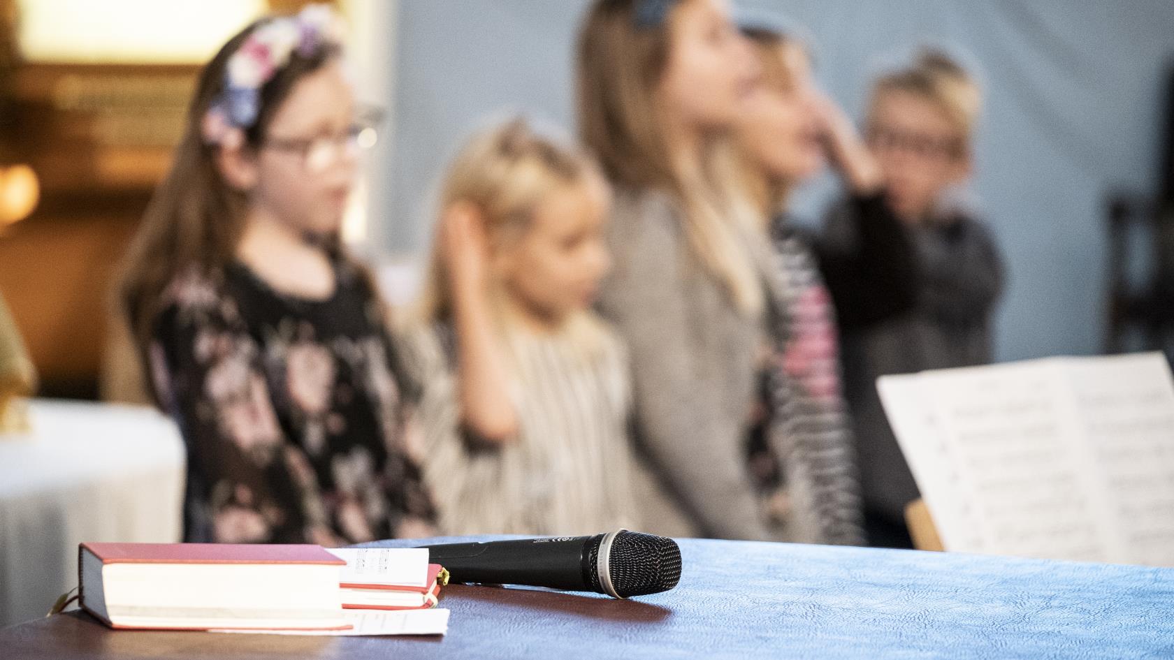 En psalmbok och en mikrofon ligger framlagda. I bakgrunden syns sjungande barn.