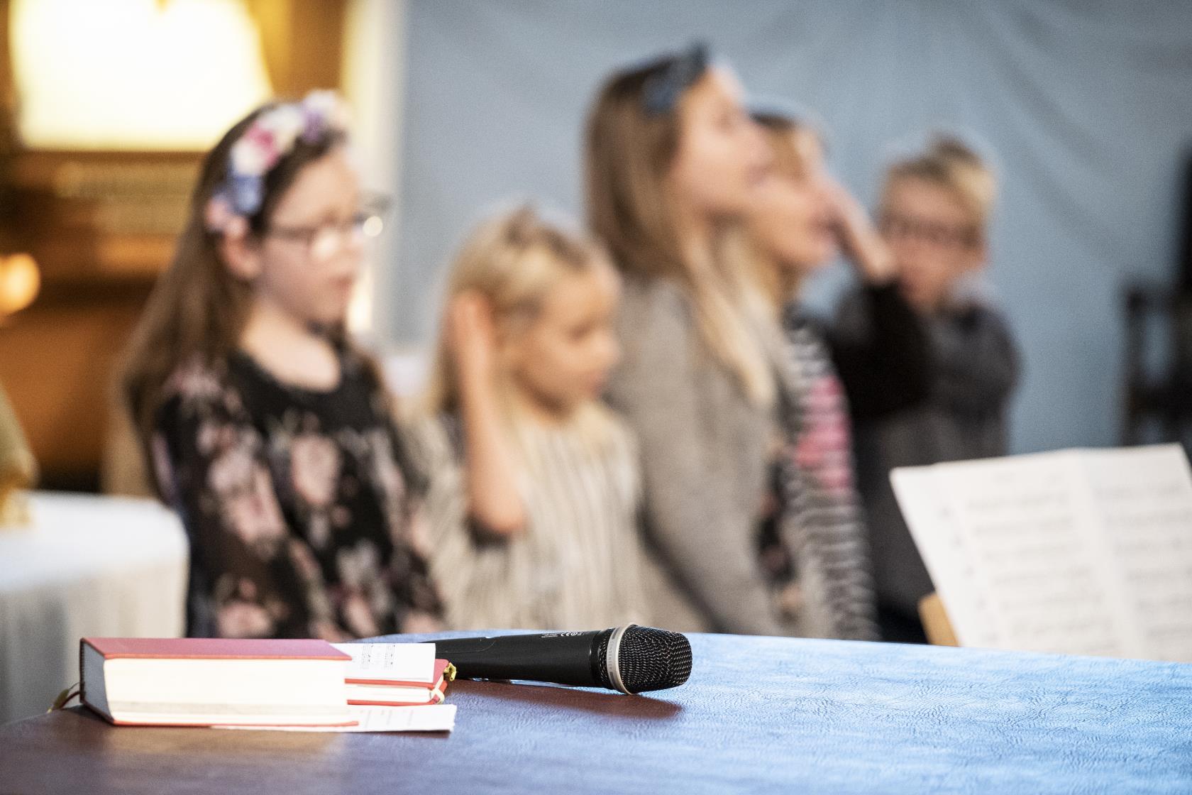 En psalmbok och en mikrofon ligger framlagda. I bakgrunden syns sjungande barn.