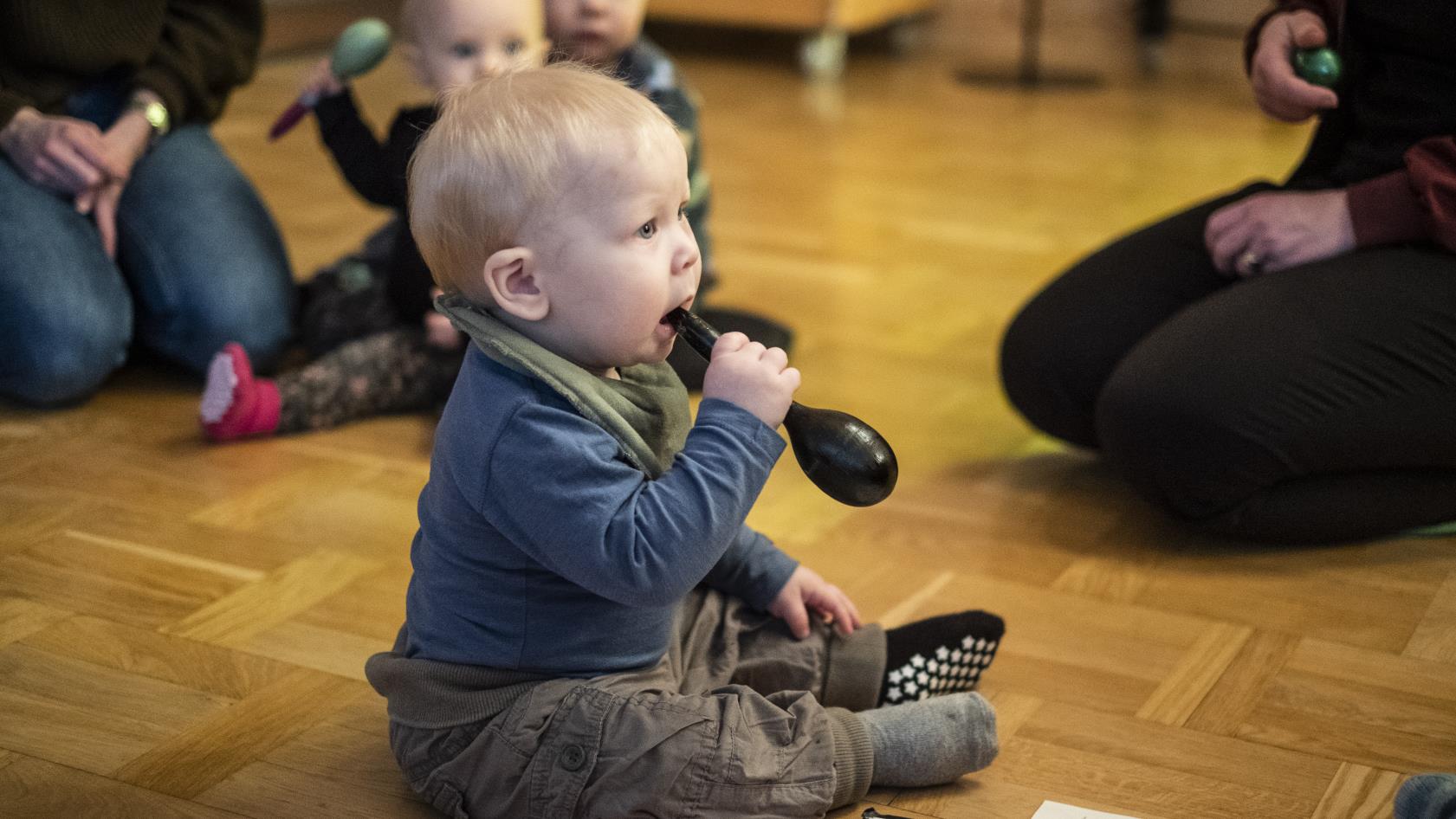 Några mammor sitter i en ring på golvet med sina bebisar. En bebis har en maracas i munnen.