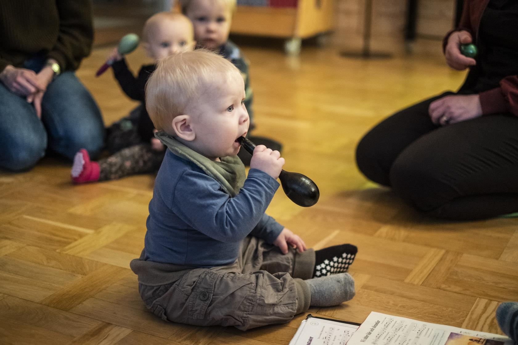 Några mammor sitter i en ring på golvet med sina bebisar. En bebis har en maracas i munnen.