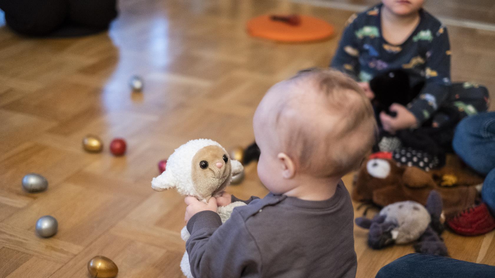 En bebis på golvet tittar på ett gosedjur som föreställer ett får.