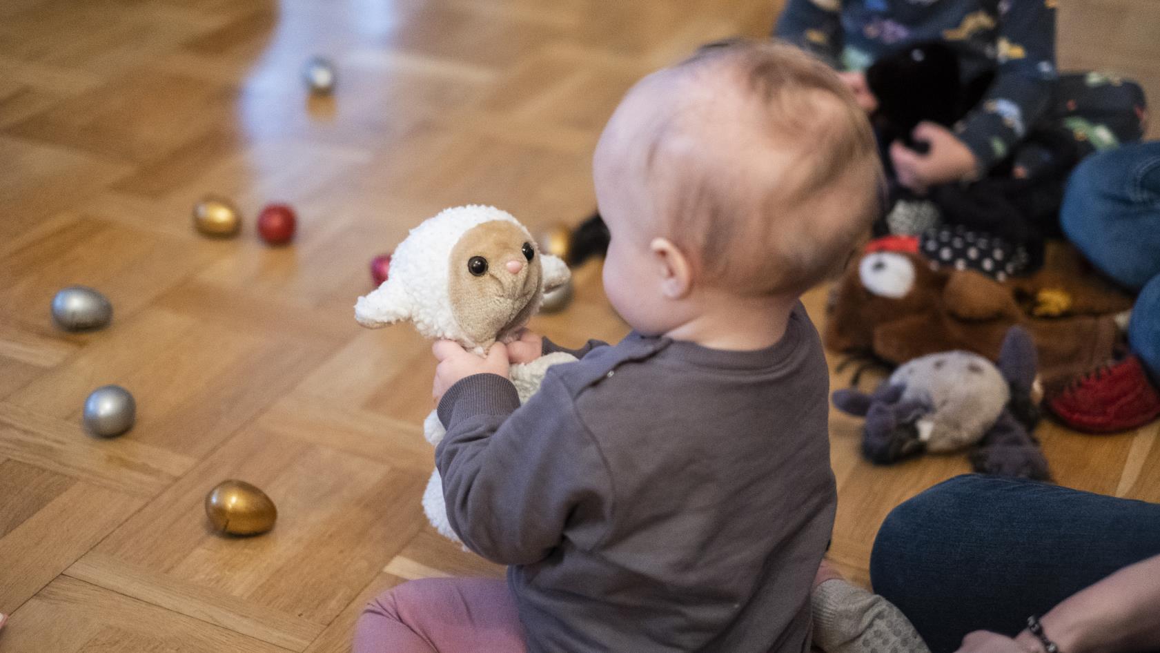 En bebis på golvet tittar på ett gosedjur som föreställer ett får.
