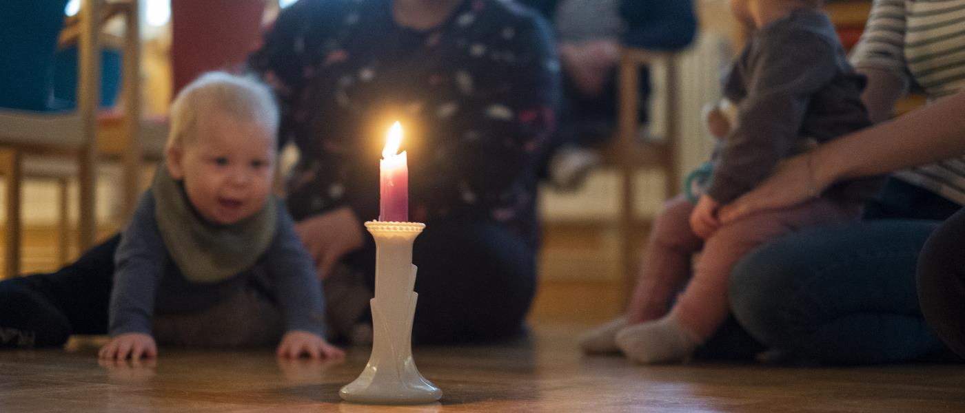 Några mammor sitter i en ring på golvet med sina bebisar. En bebis tittar nyfiket på ett levande ljus som står i mitten.