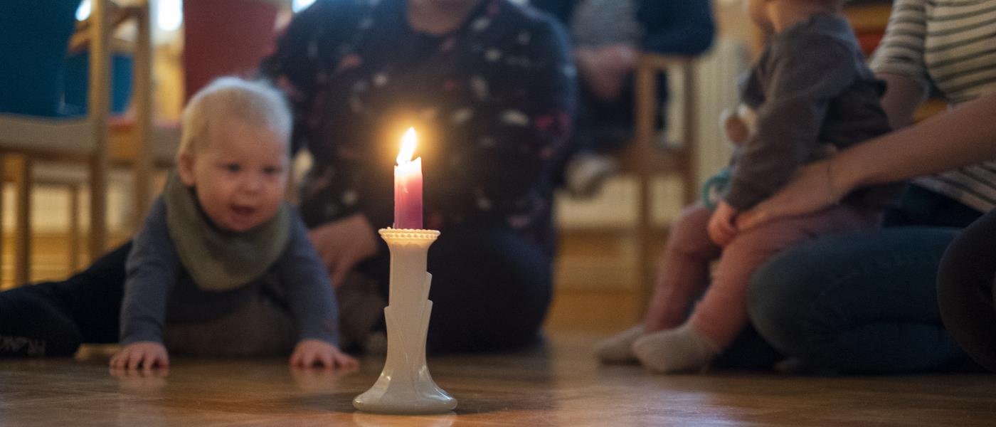 Några mammor sitter i en ring på golvet med sina bebisar. En bebis tittar nyfiket på ett levande ljus som står i mitten.