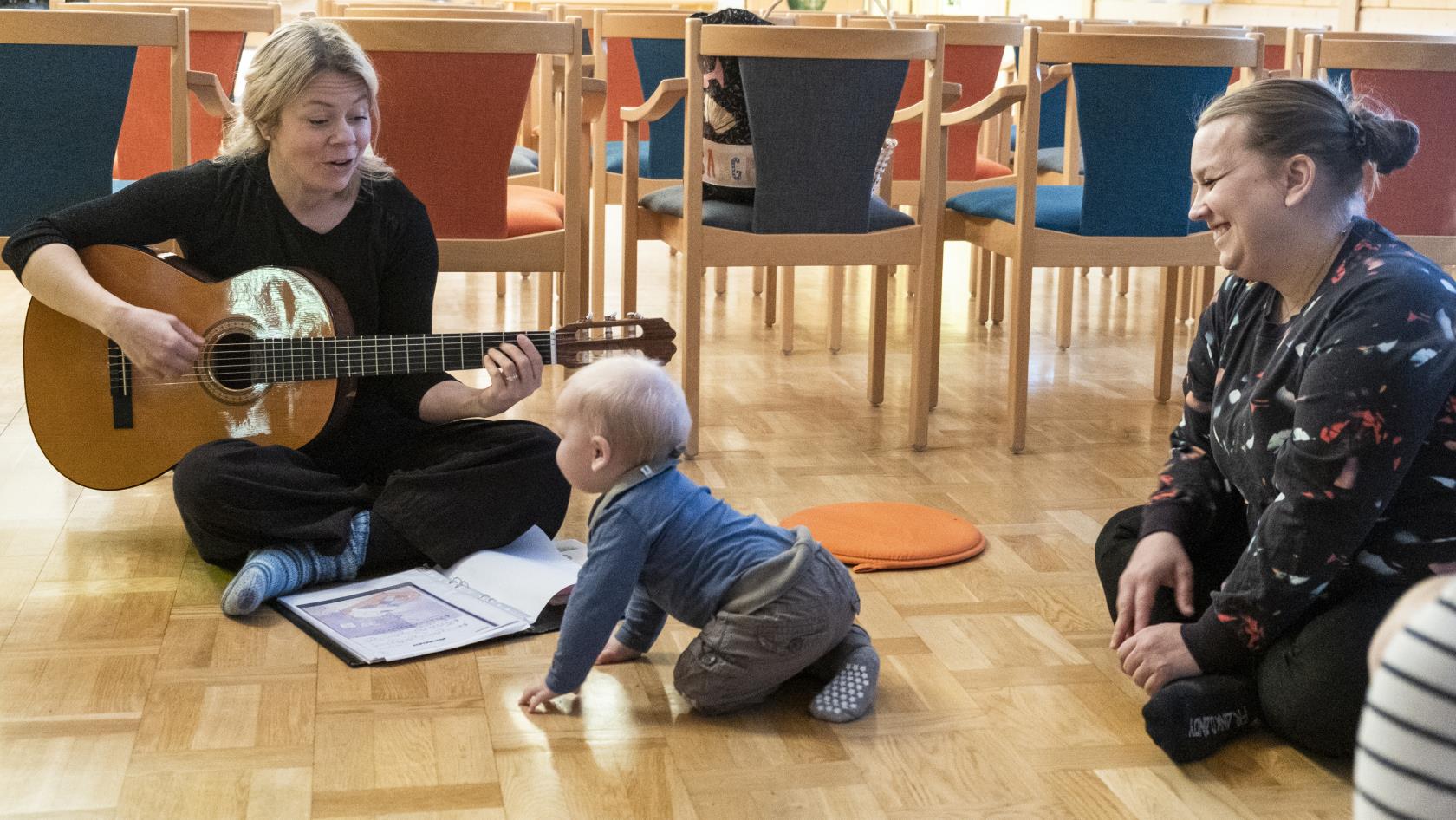 Ett litet barn kryper fram mot fröken som sitter i en ring på golvet och sjunger och spelar gitarr på Öppen förskola. Barnets mor ser på.