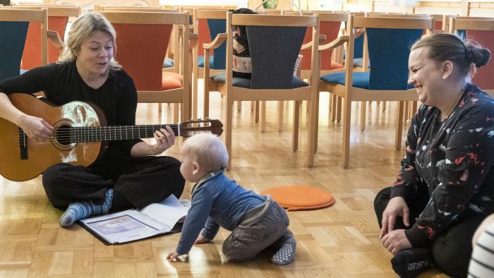 Ett litet barn kryper fram mot fröken som sitter i en ring på golvet och sjunger och spelar gitarr på Öppen förskola. Barnets mor ser på.