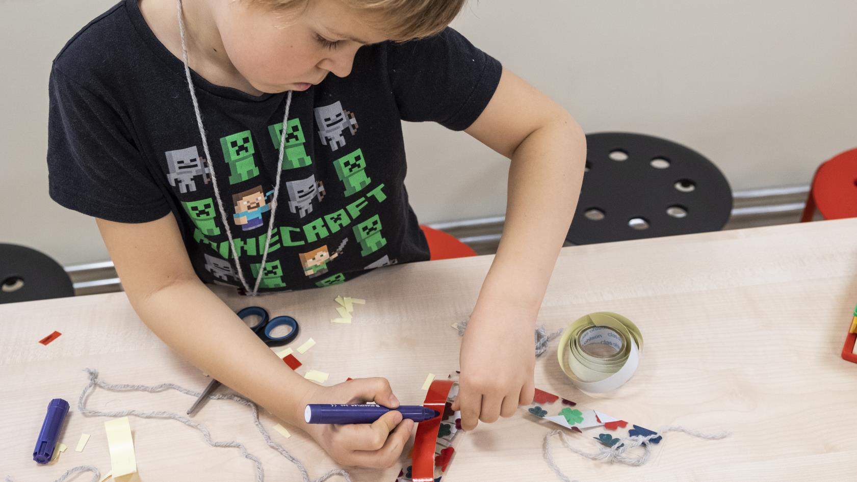 En pojke i lågstadieålder pysslar koncentrerat med en blå tuschpenna, rött glansigt papper. Pojken bär en svart T-shirt  med Minecraftfigurer.