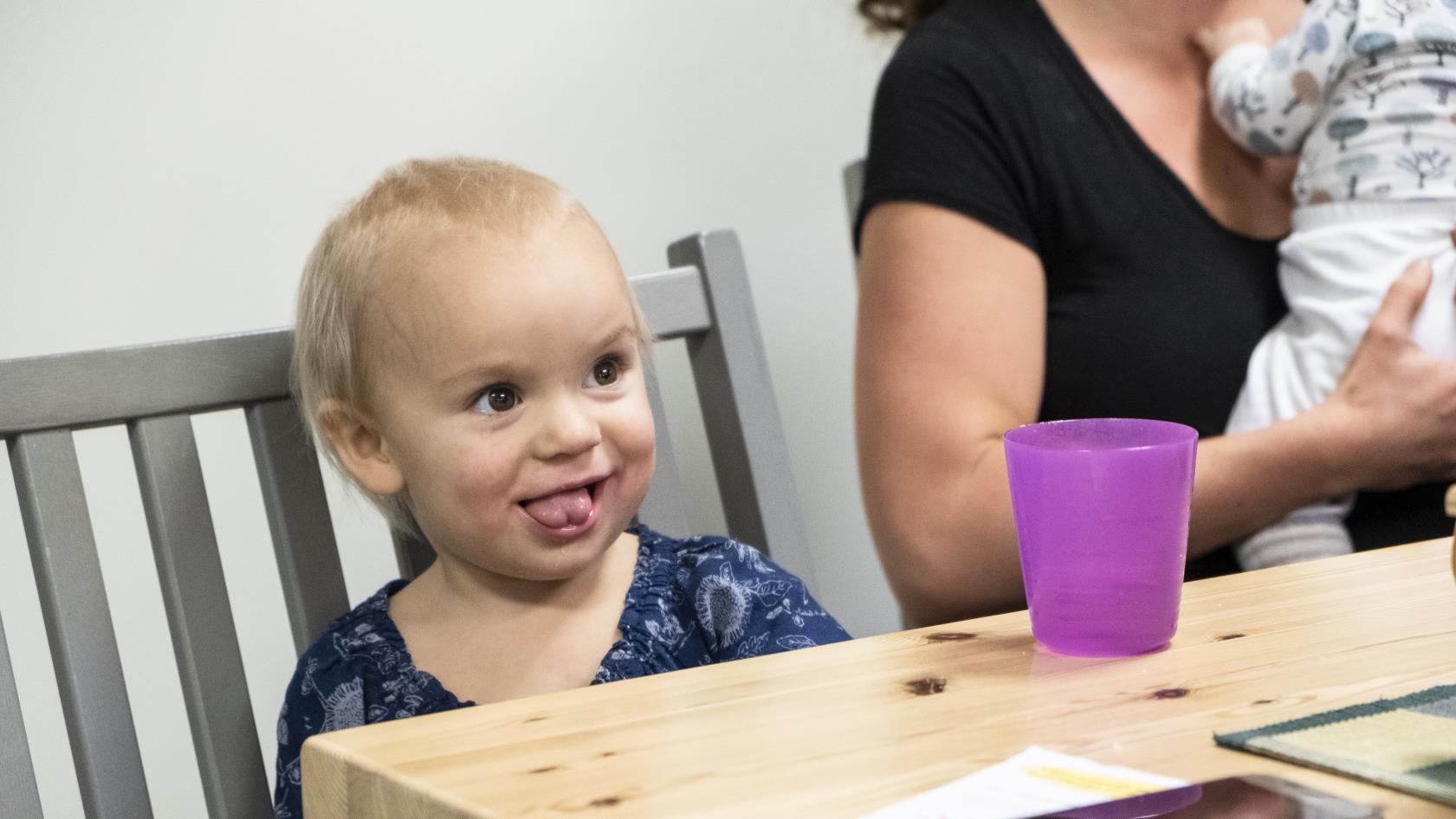 Ett barn på några år sitter vid ett bord på en grå pinnstol. På bordet står en lila plastmugg. I bakgrunden anar vi en amma med ett spädbarn.