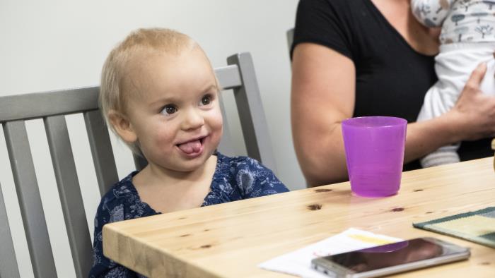 Ett barn på några år sitter vid ett bord på en grå pinnstol. På bordet står en lila plastmugg. I bakgrunden anar vi en amma med ett spädbarn.