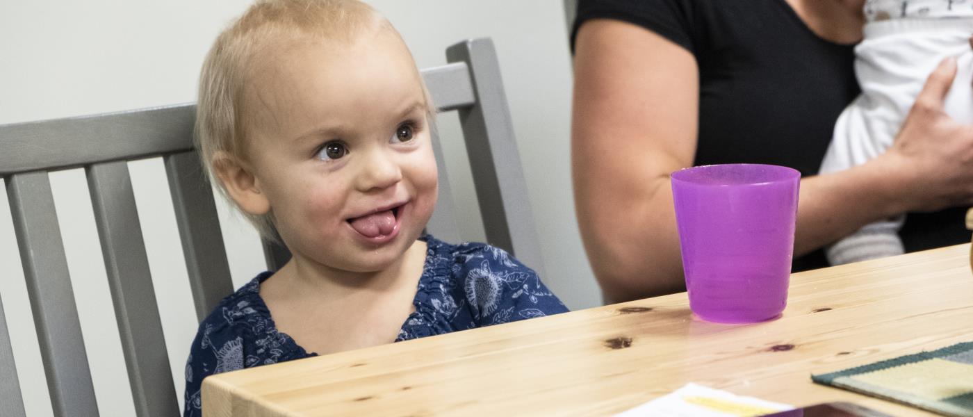 Ett barn på några år sitter vid ett bord på en grå pinnstol. På bordet står en lila plastmugg. I bakgrunden anar vi en amma med ett spädbarn.