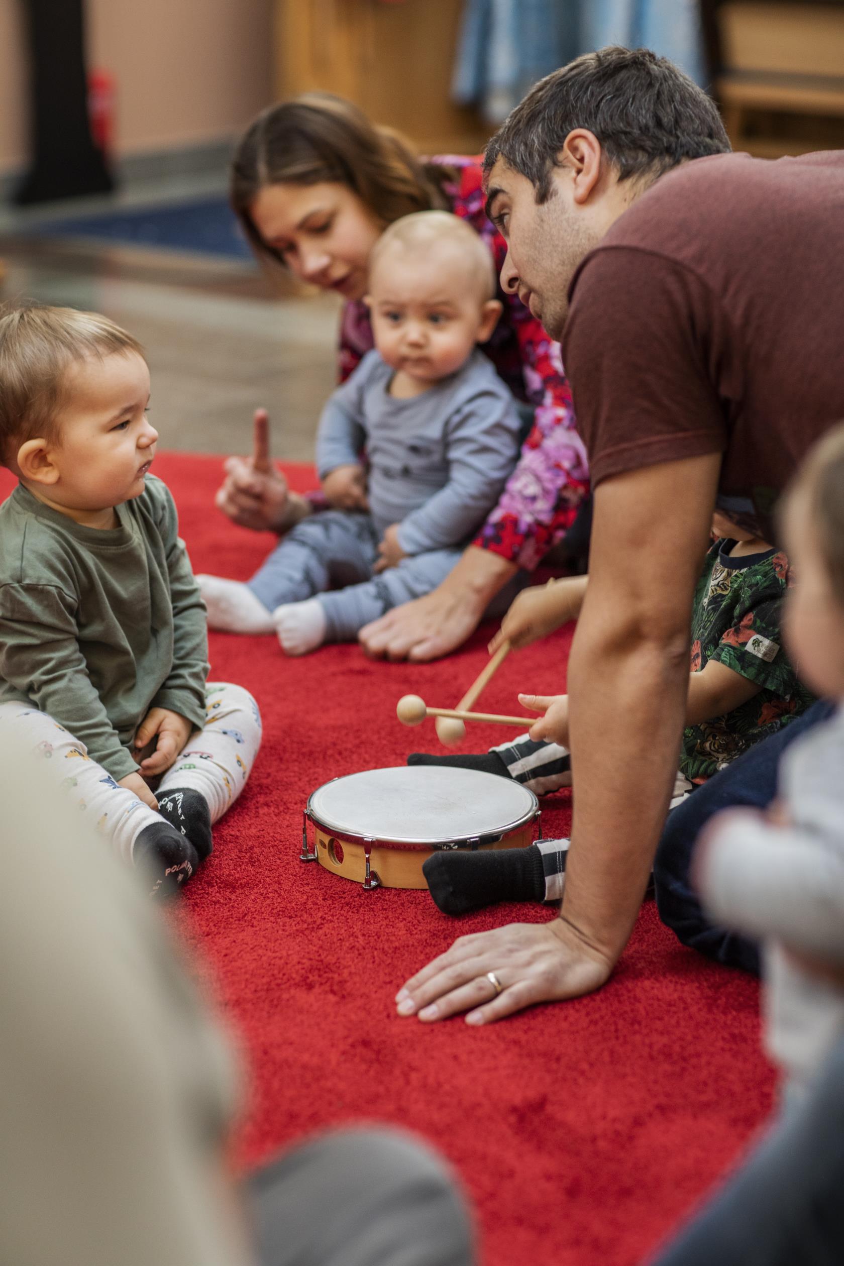 Ett litet barn spelar på en tamburin som ligger på en röd matta. Andra barn och vuxna sitter runt omkring.