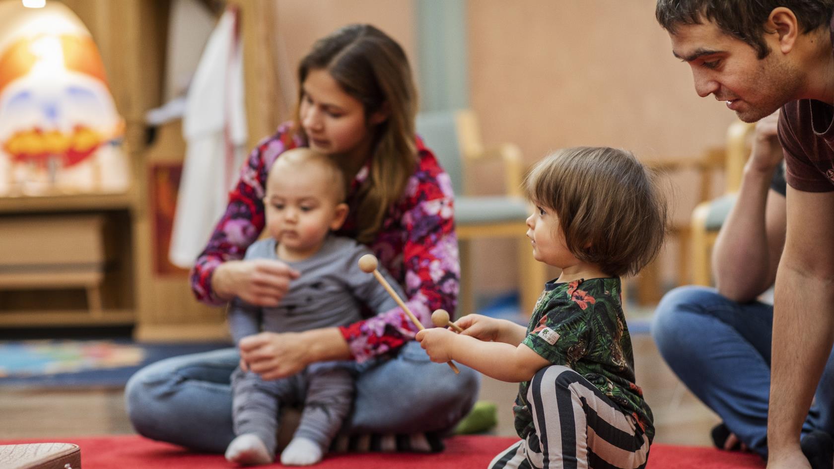 Ett litet barn spelar på en tamburin som ligger på en röd matta. Andra barn och vuxna sitter runt omkring.