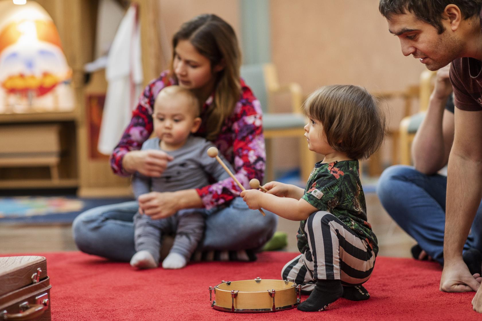 Ett litet barn spelar på en tamburin som ligger på en röd matta. Andra barn och vuxna sitter runt omkring.