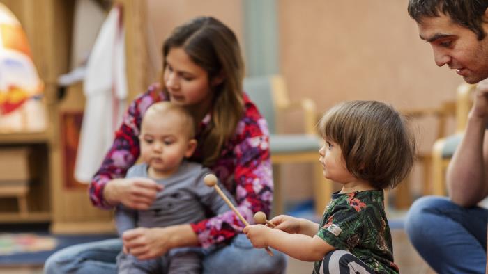 Ett litet barn spelar på en tamburin som ligger på en röd matta. Andra barn och vuxna sitter runt omkring.