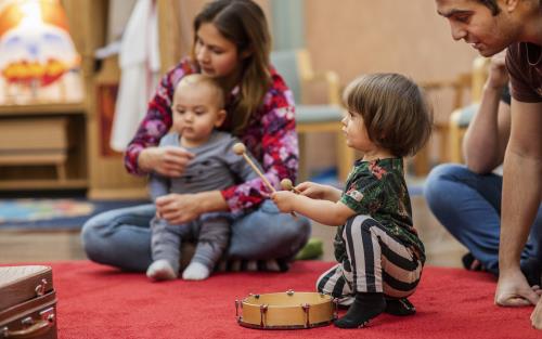 Ett litet barn spelar på en tamburin som ligger på en röd matta. Andra barn och vuxna sitter runt omkring.