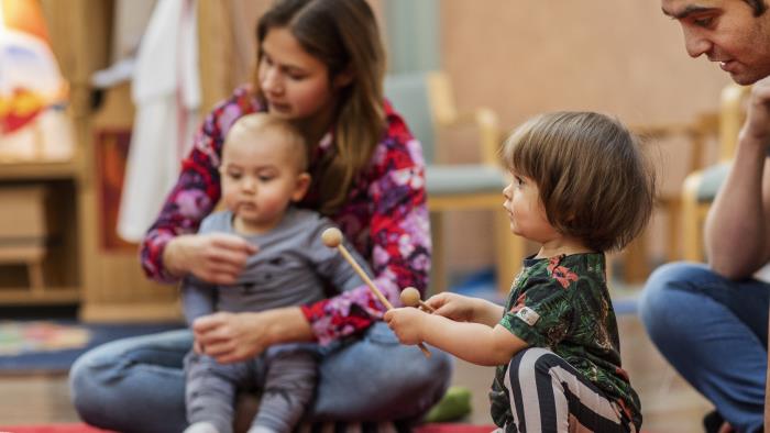 Ett litet barn spelar på en tamburin som ligger på en röd matta. Andra barn och vuxna sitter runt omkring.