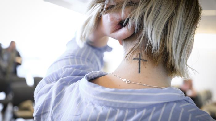 En kvinna lyfter upp sitt blonda hår i nacken för att visa en tatuering som föreställer ett kors.
