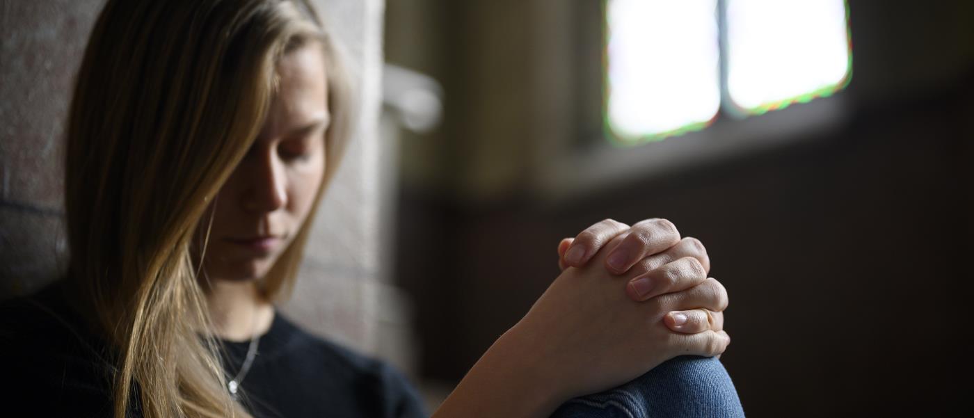 En ung kvinna med knäppta händer och slutna ögon sitter mot en stenvägg i en kyrka.