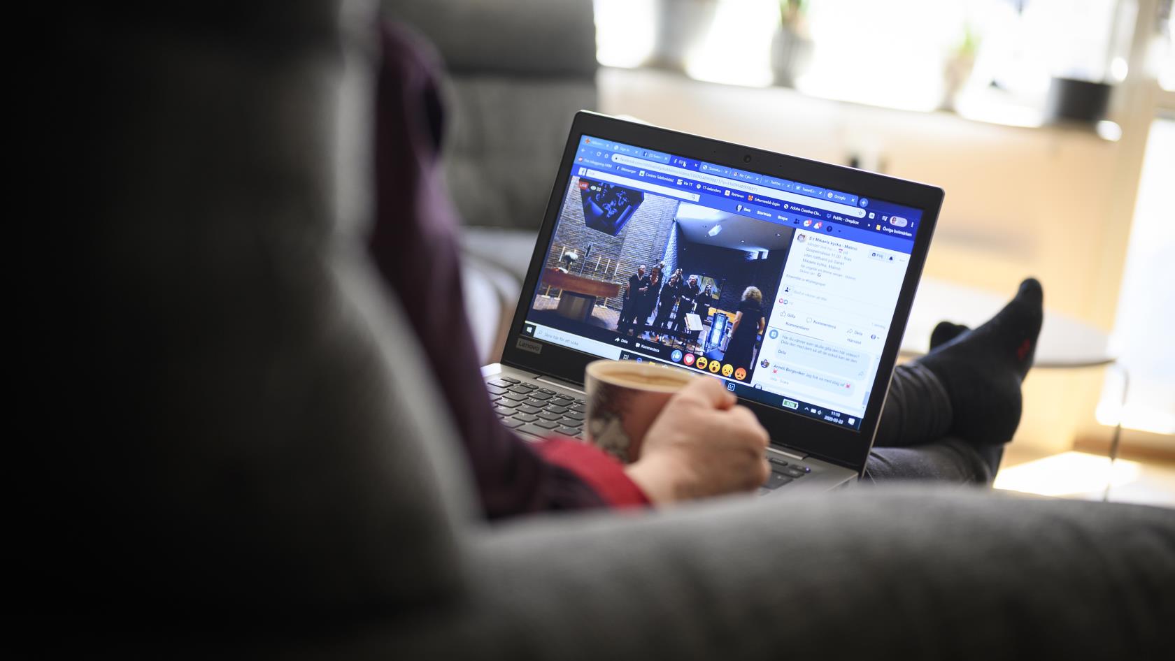 En person sitter i en soffa med sin dator i knät och följer en digital gudstjänst.