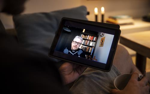 En person sitter med en läsplatta i knät. På skärmen syns en präst i hemmiljö.