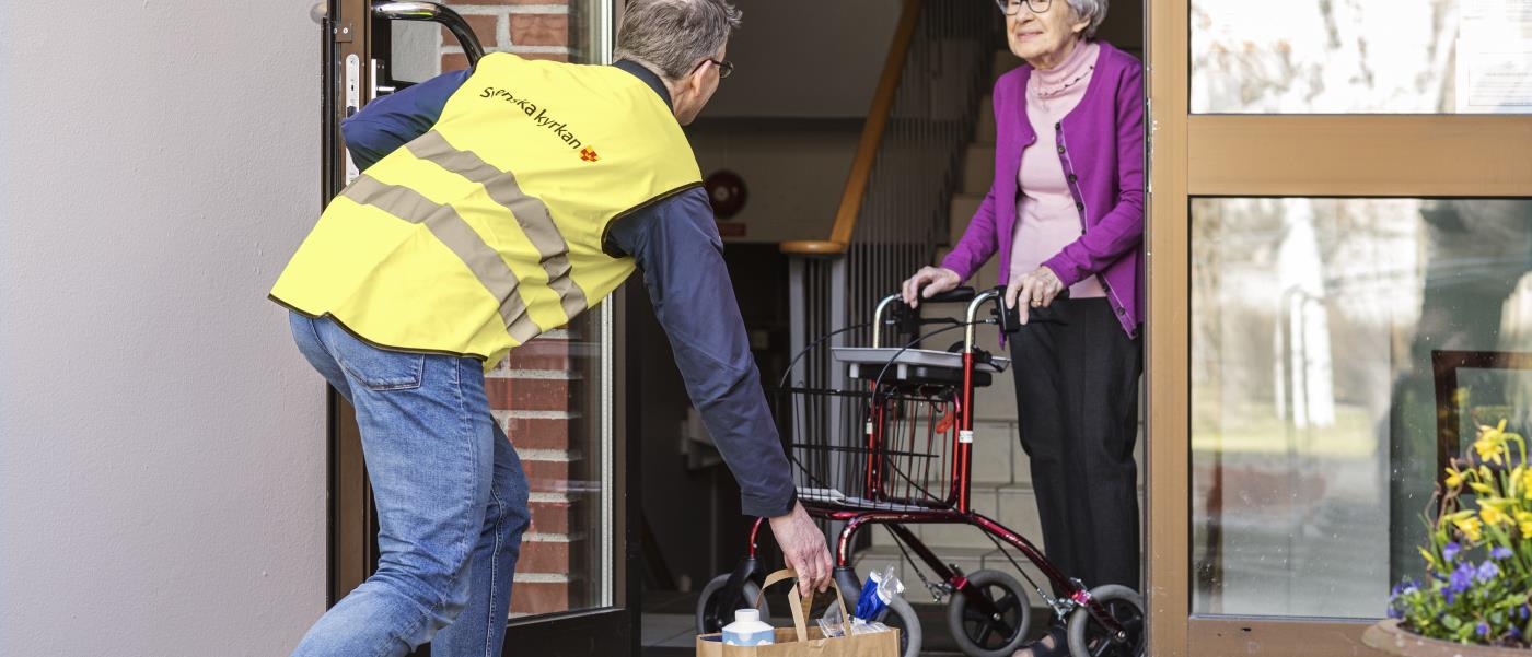 En man från Svenska kyrkan lämnar en matkasse till en äldre dam vid ytterdörren.