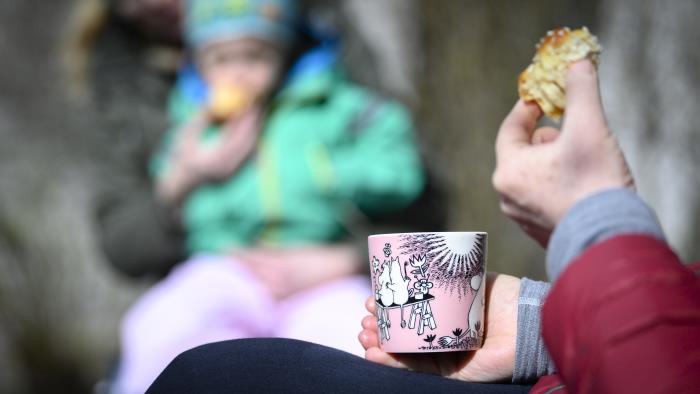 Någon sitter och fikar kaffe och bulle. I bakgrunden sitter ett barn i knät på en vuxen.