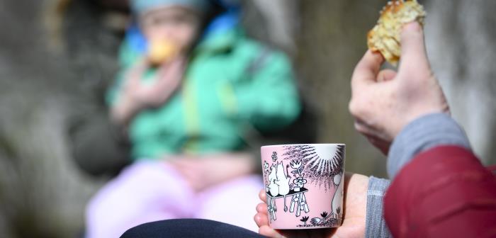 Någon sitter och fikar kaffe och bulle. I bakgrunden sitter ett barn i knät på en vuxen.
