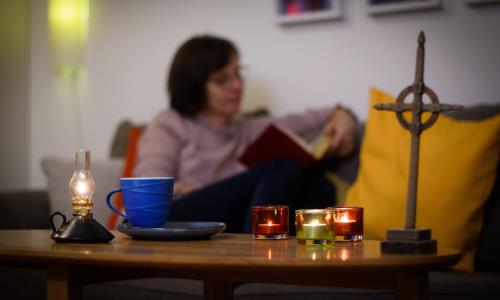 En kvinna läser hemma i soffan. På soffbordet framför henne står tända ljus och ett kors.