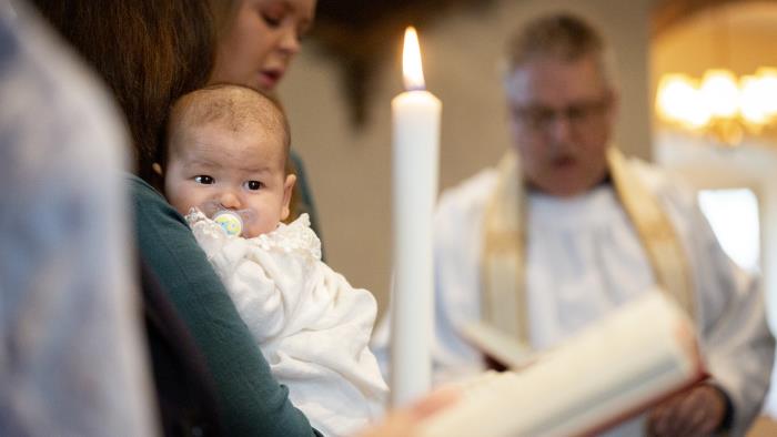 En bebis i dopklänning vilar i någons famn. I bakgrunden står en präst.