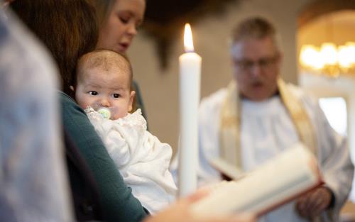 En bebis i dopklänning vilar i någons famn. I bakgrunden står en präst.