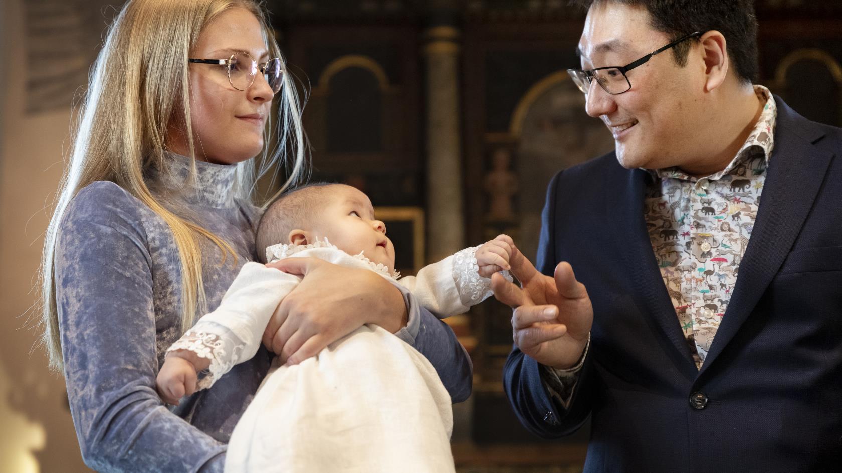 En mor står med sitt lilla barn i famnen i kyrkan. Barnet tittat glatt mot sin far som håller i dess hand.