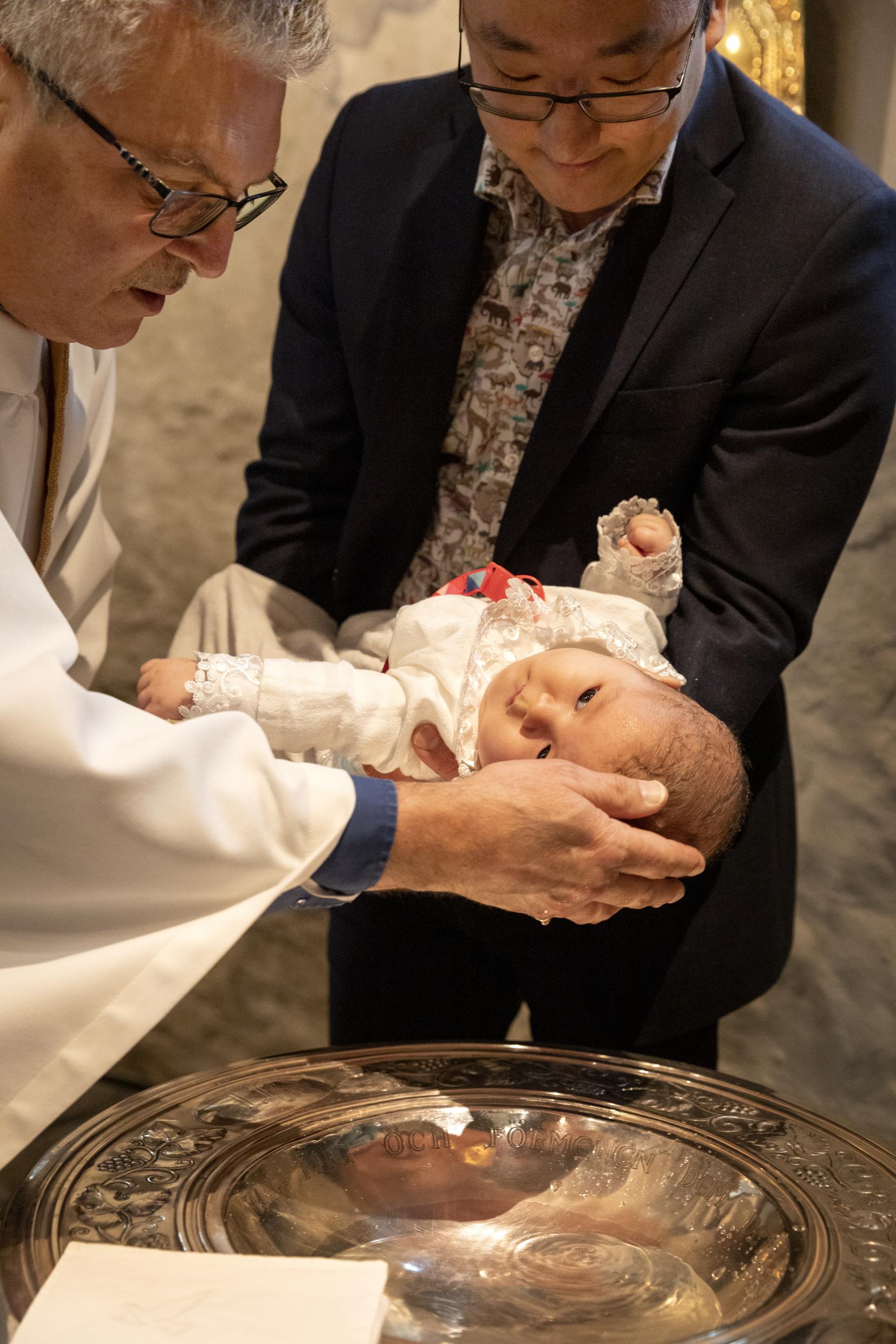En bebis i dopklänning får vatten på sitt huvud av en präst.