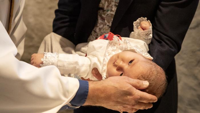 En bebis i dopklänning får vatten på sitt huvud av en präst.