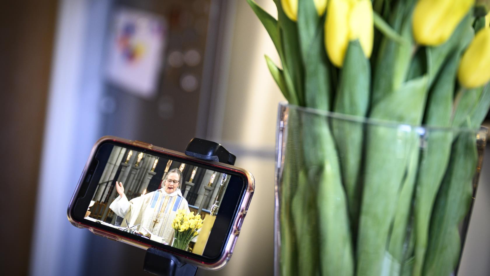 En mobiltelefon står uppställd bredvid en vas tulpaner. Skärmen visar en biskop som håller gudstjänst.