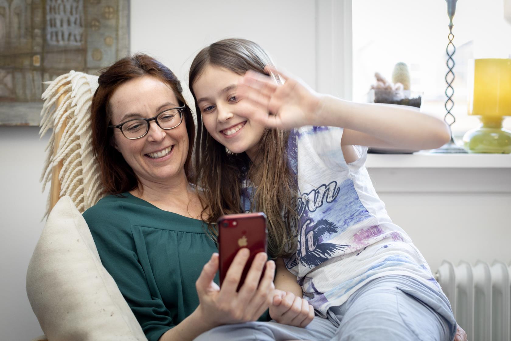 En mamma med sin dotter i knät vinkar åt någon som de videosamtalar med i mobiltelefonen.