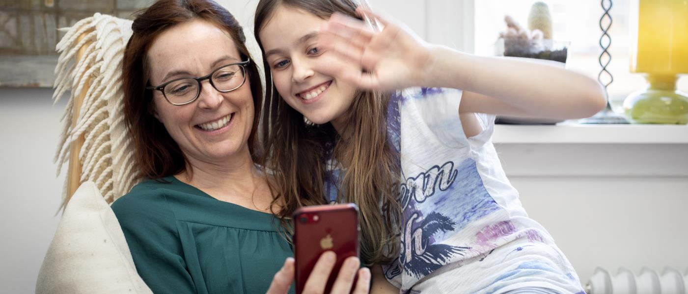 En mamma med sin dotter i knät vinkar åt någon som de videosamtalar med i mobiltelefonen.