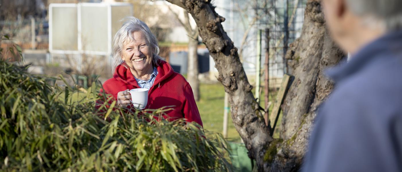 Leende kvinna med kaffekopp i handen står i sin trädgård och pratar med granne på andra sidan häcken.
