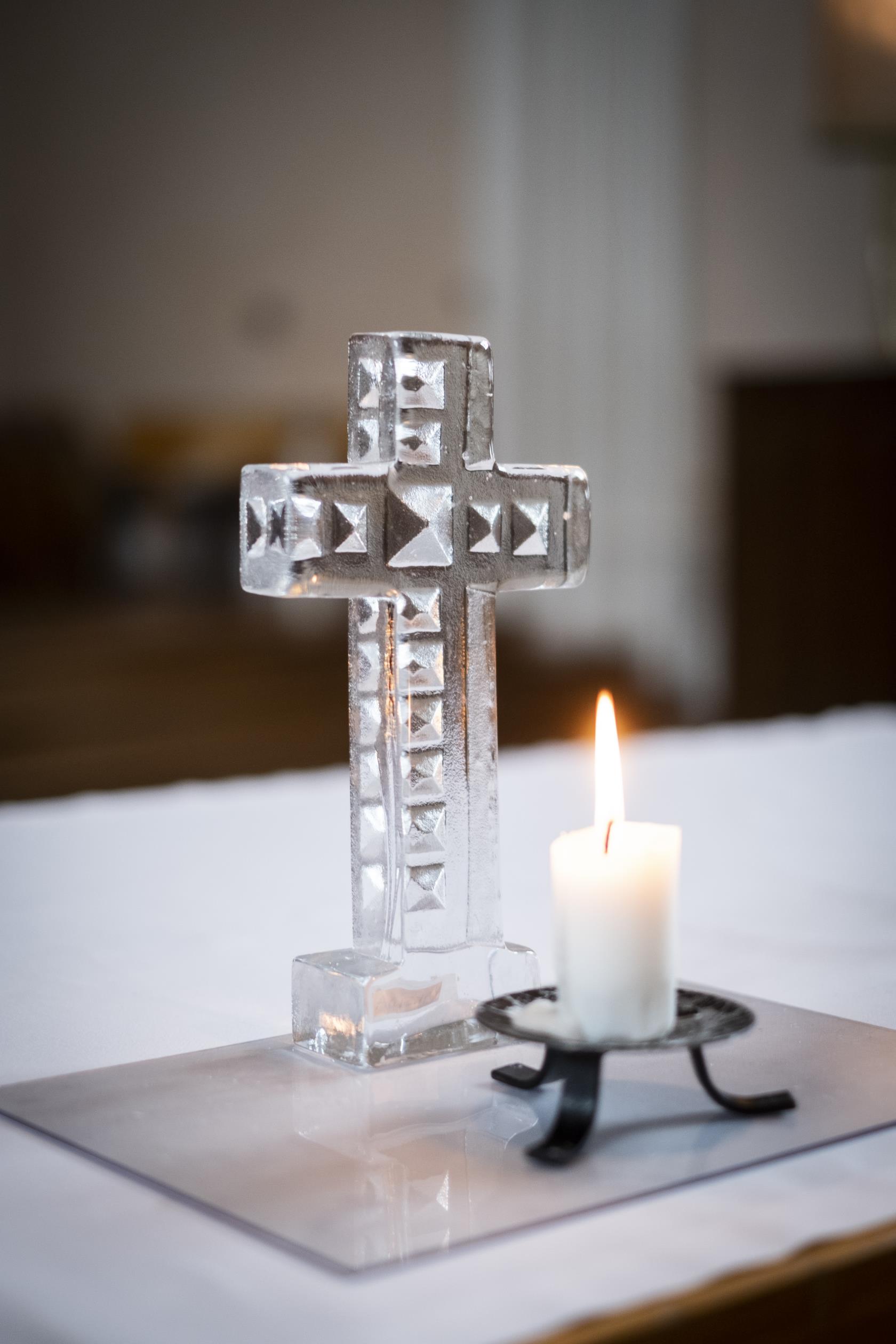 Ett kors av handgjort glas står på ett altare. Framför korset brinner ett ljus på en trefotad ljusstake.