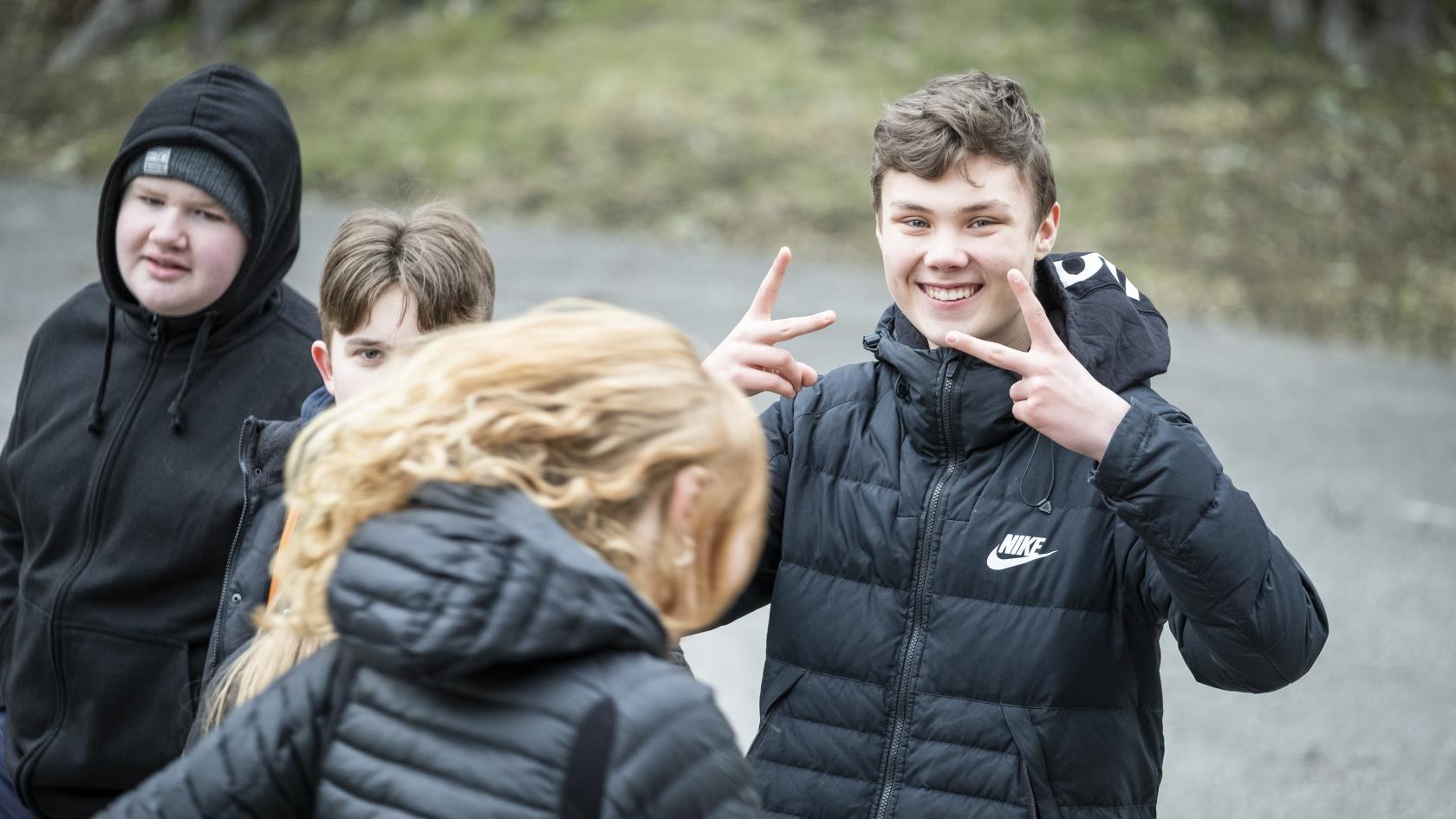 En ung kille ler mot kameran och gör peace-tecken med händerna. Hans kompisar står bredvid.