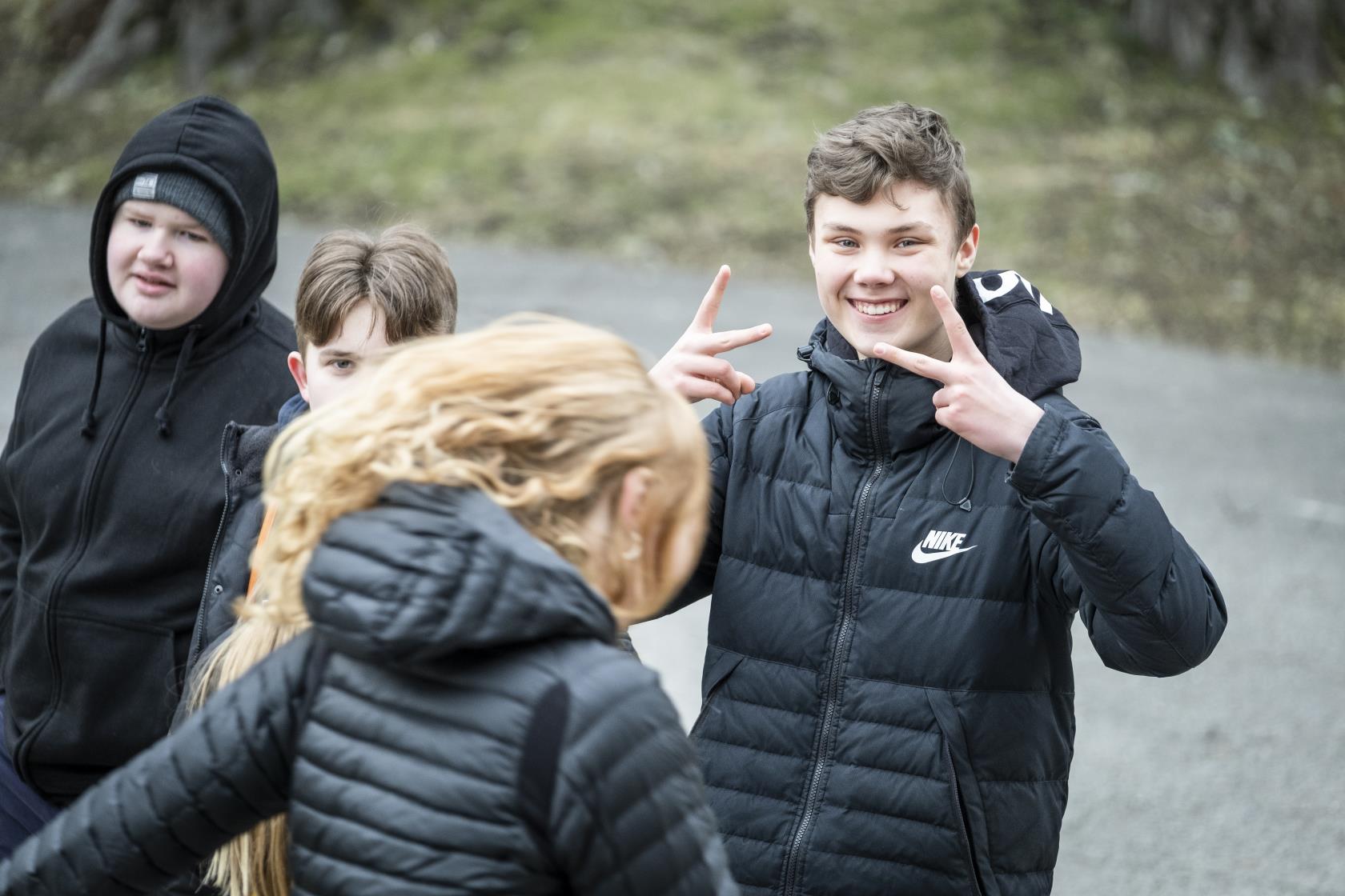 En ung kille ler mot kameran och gör peace-tecken med händerna. Hans kompisar står bredvid.