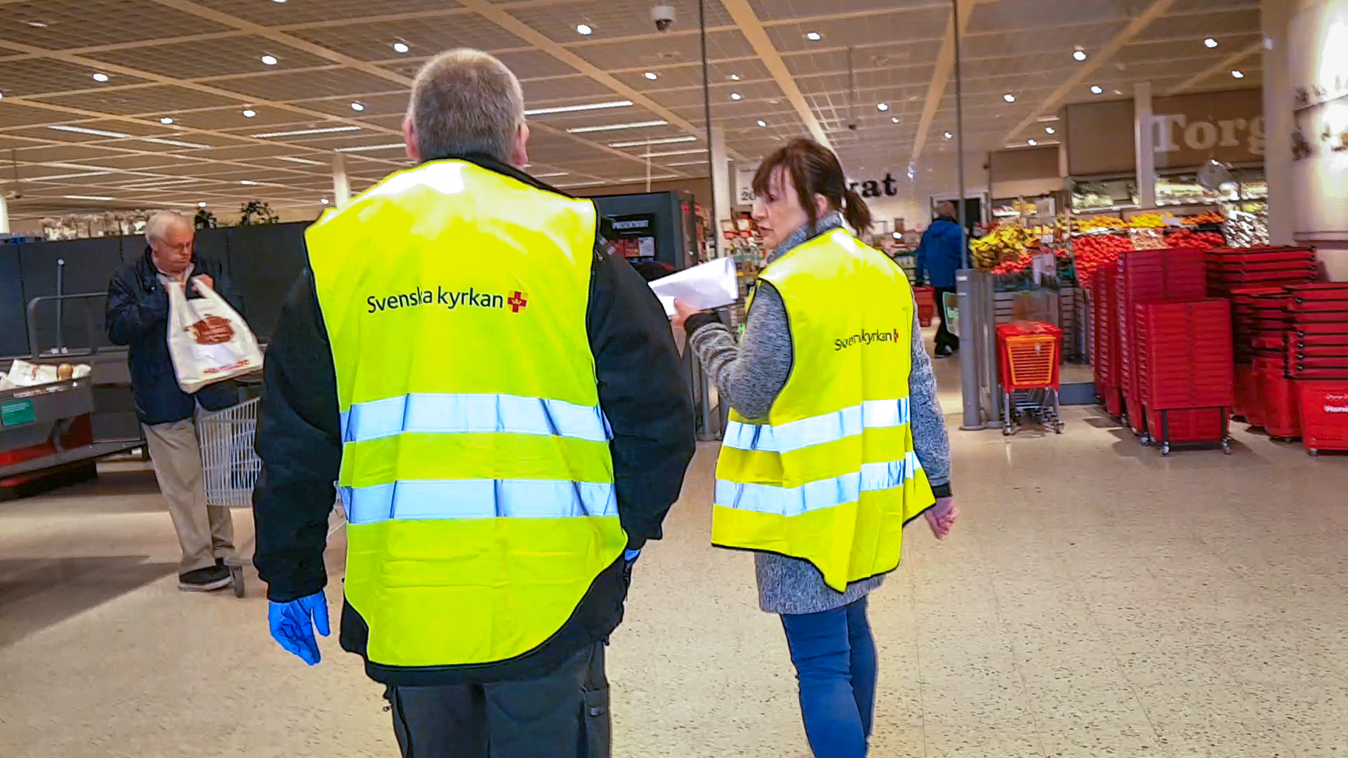 Två personer i Svenska kyrkans reflexvästar är på väg in i en matbutik.