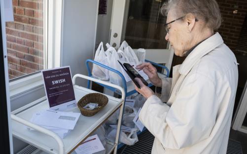 En äldre dam står med mobiltelefonen framför en skylt med texten Här kan du använda Swish.