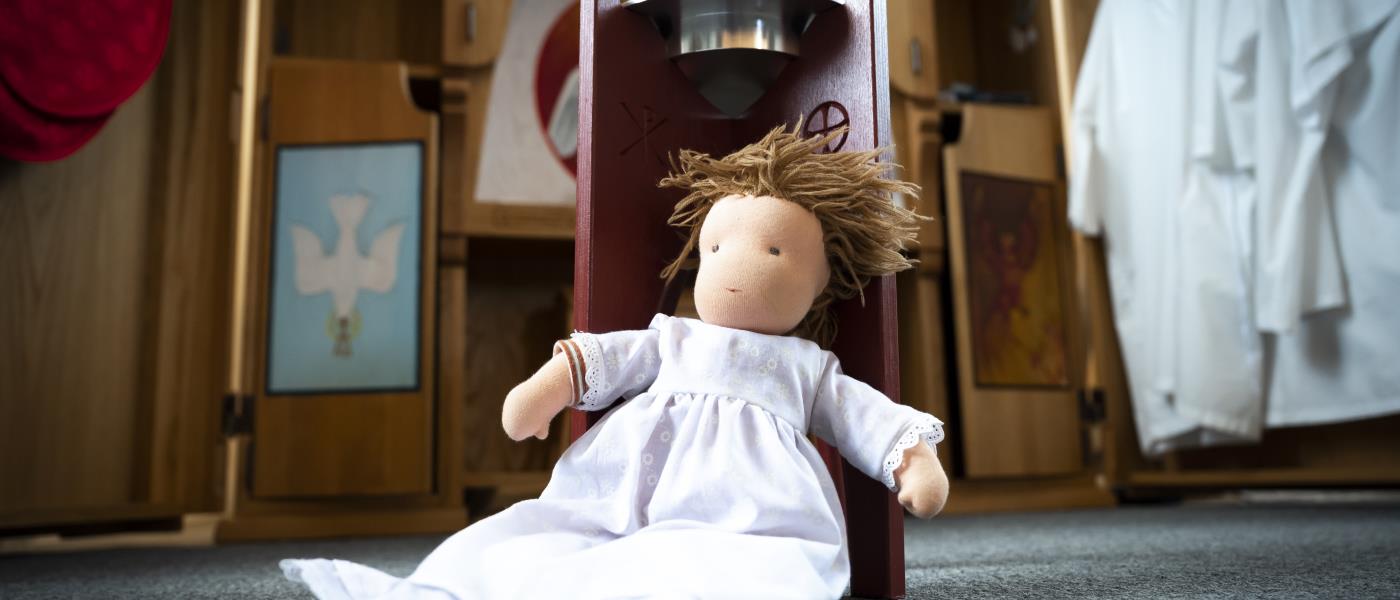 En docka i dopklänning sitter lutad mot dopfunten.