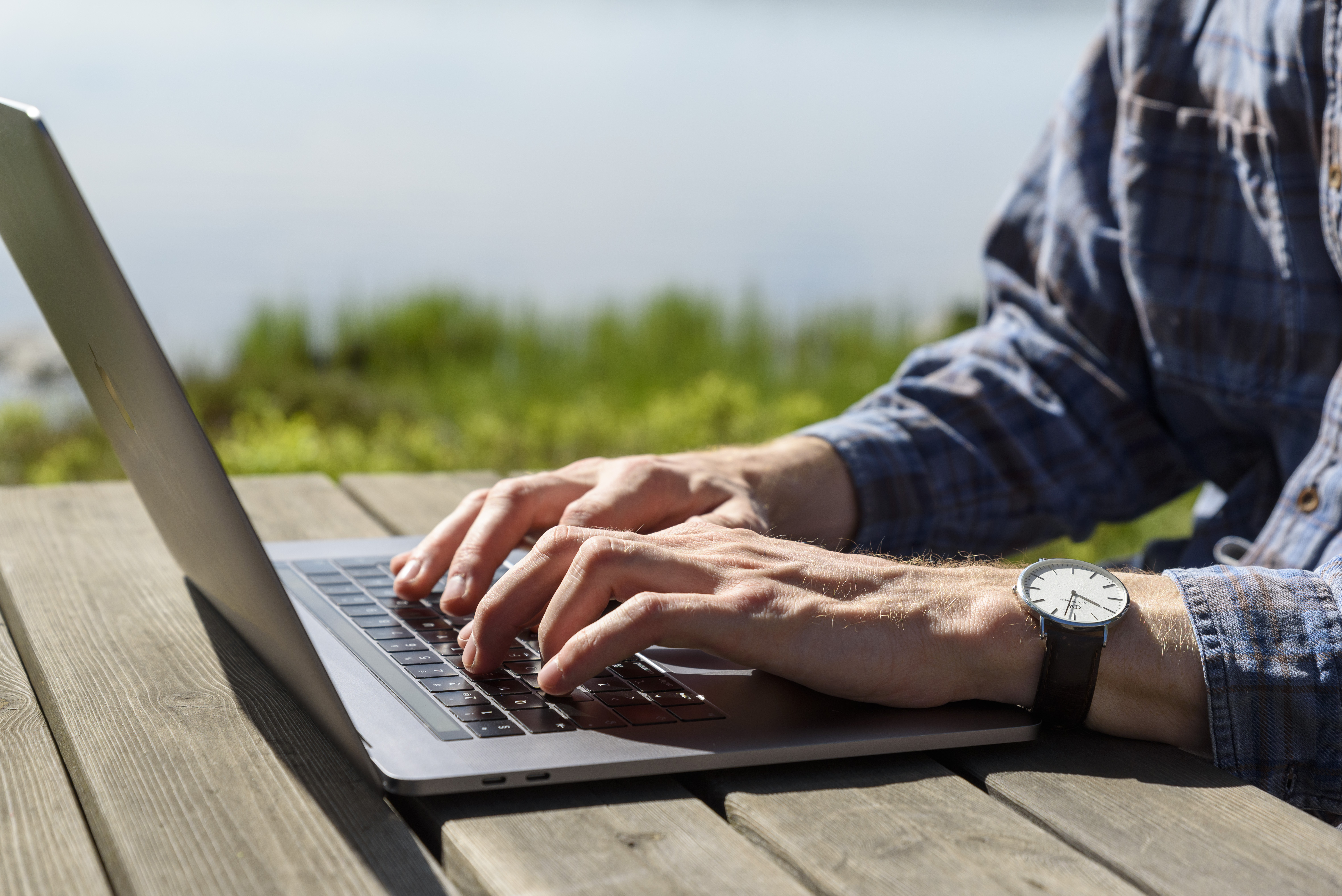 En person sitter vid ett träbord vid en sjö och jobbar på sin laptop.