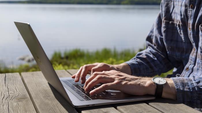 En person sitter vid ett träbord vid en sjö och jobbar på sin laptop.